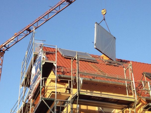 Kran hebt Solarpanele über das Dach eines Gebäudes. Das Bild zeigt das Dach und die Installation der Solarpanele.